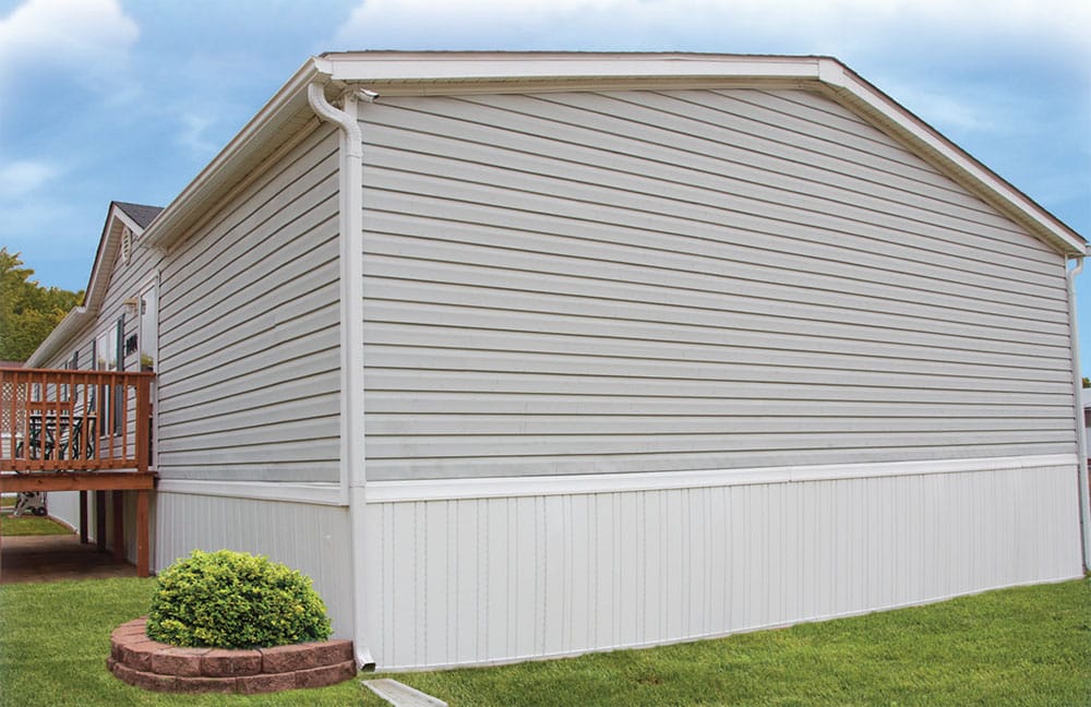 A light-colored mobile home with a wooden deck and small bush on grass. The sky is partly cloudy, no people are visible.