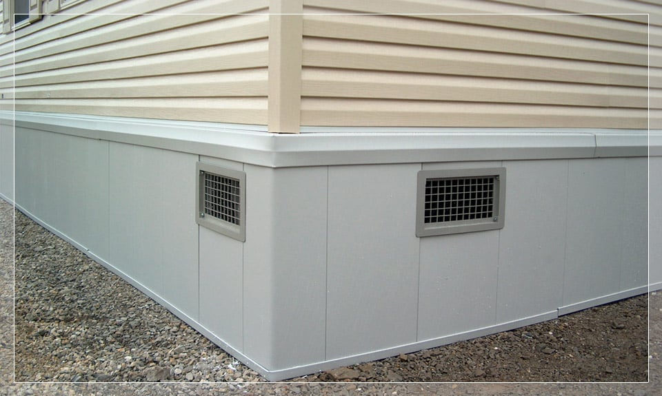 Exterior foundation of a building with beige siding and white skirting, featuring two vent openings, with a gravel base surrounding the structure.