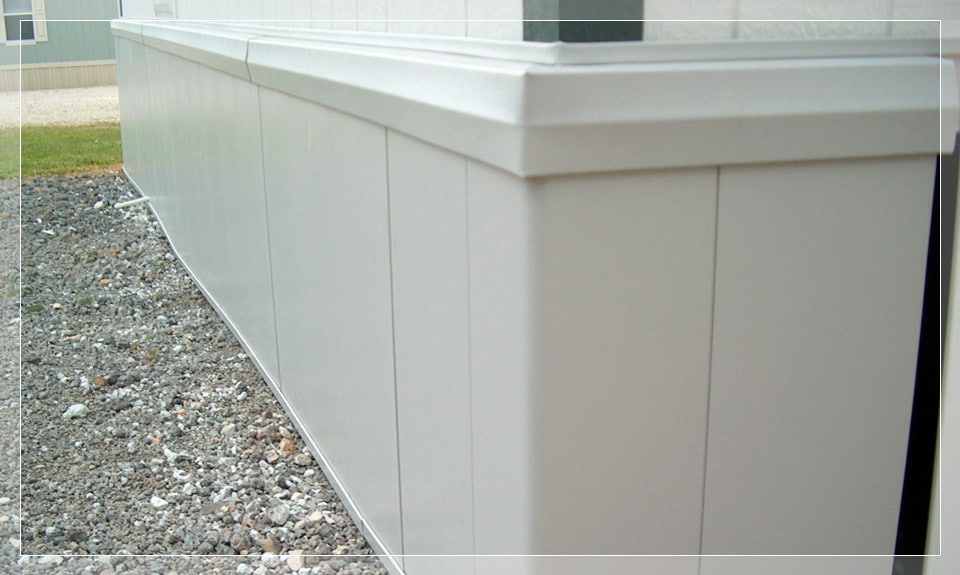 The image shows a close-up of a white vinyl skirting on a manufactured home, with gravel at the base and a grassy area.
