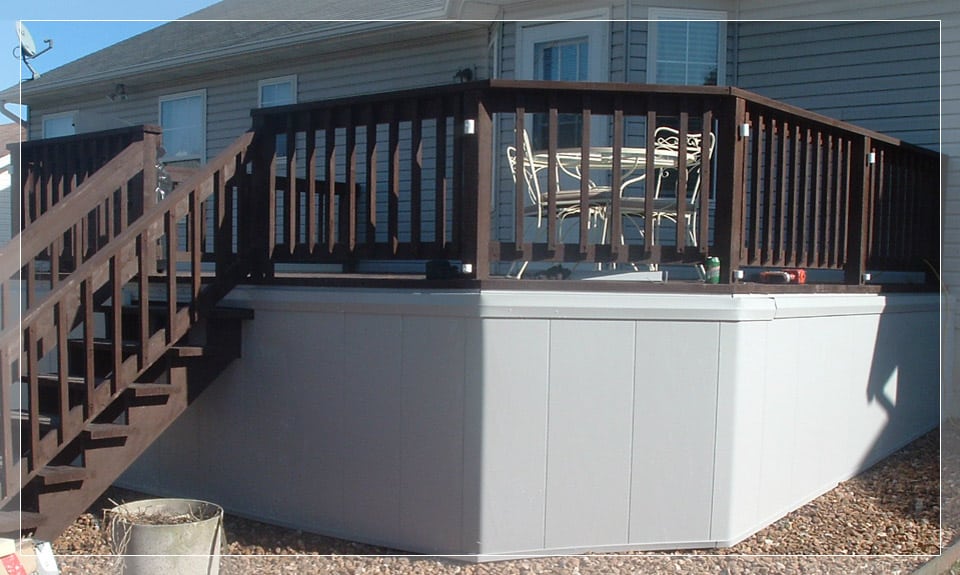 A wooden deck with railing surrounds an elevated above-ground pool, adjacent to a house with gray siding and windows. Outdoor furniture is visible.