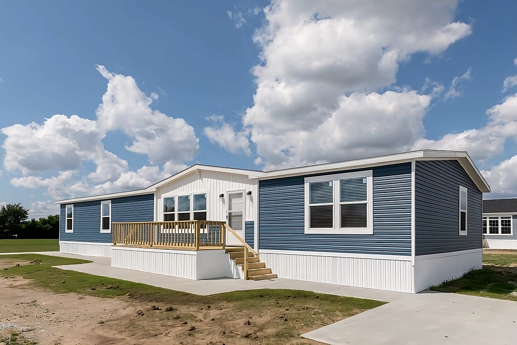 A modern blue manufactured home with a small wooden porch sits under a partly cloudy sky, surrounded by a grass and dirt landscape.